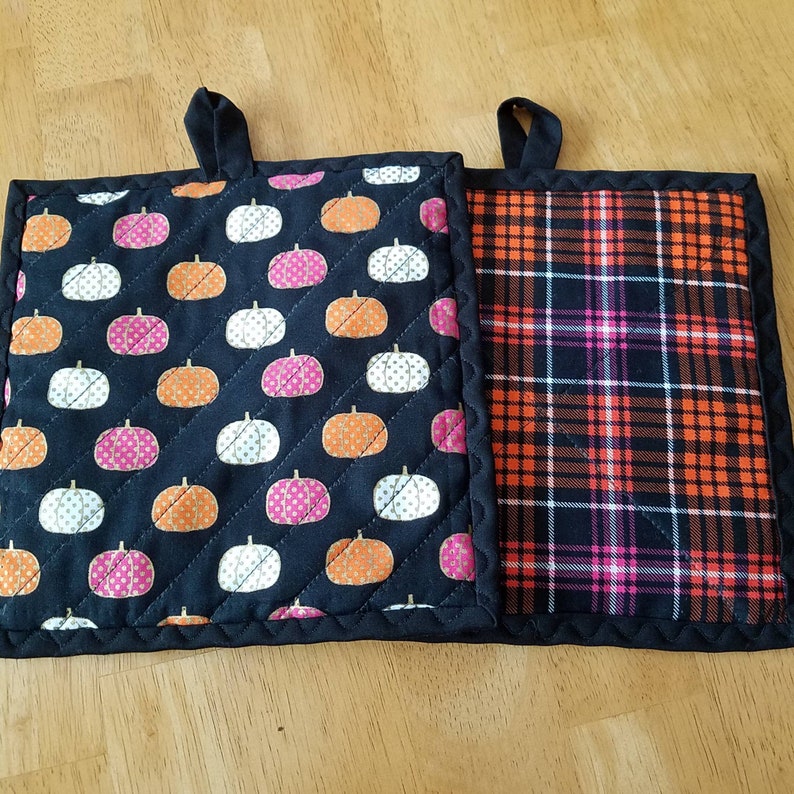 May include: Two black quilted pot holders. One has a black and white pumpkin pattern and the other has a black, orange, pink, and white plaid pattern.