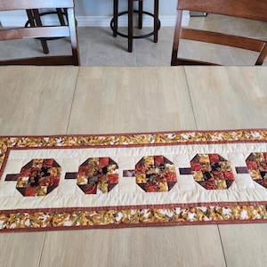 以下が含まれることがあります： クリーム色の中心部と秋の葉の縁取りが付いた、長方形のキルトテーブルランナー。中央には、赤、茶色、オレンジの色合いの5つの八角形の形があり、茶色のバーで接続されています。 ランナーは明るい色の木製のテーブルの上に置かれています。