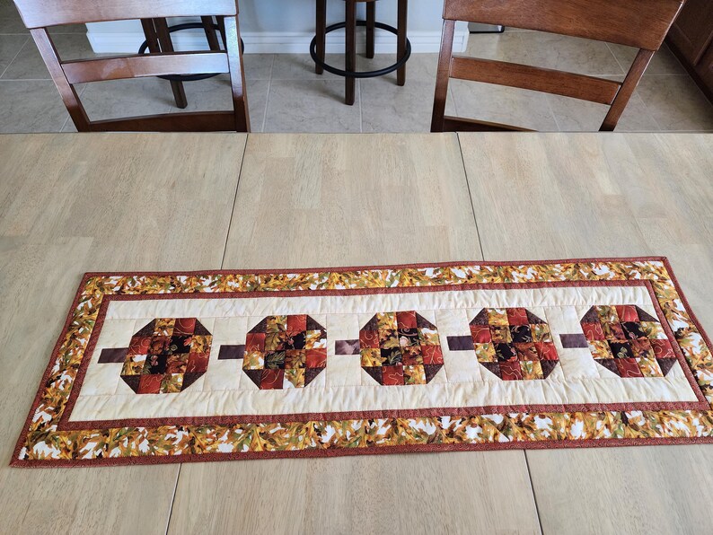 以下が含まれることがあります： クリーム色の中心部と秋の葉の縁取りが付いた、長方形のキルトテーブルランナー。中央には、赤、茶色、オレンジの色合いの5つの八角形の形があり、茶色のバーで接続されています。 ランナーは明るい色の木製のテーブルの上に置かれています。