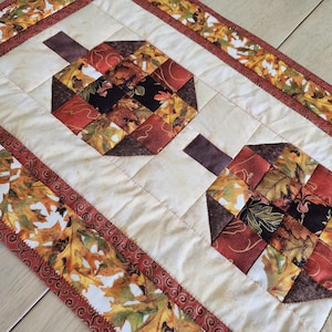 以下が含まれることがあります： 2つのカボチャのデザインが特徴のキルトテーブルランナー。カボチャは秋色のパッチワーク正方形でできており、茶色の茎が付いています。ボーダーには、秋の葉の繰り返し模様があります。