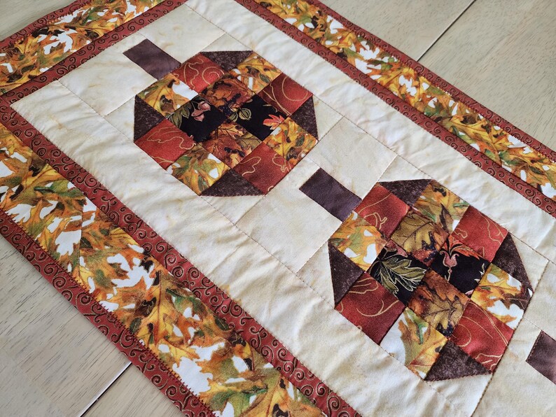 以下が含まれることがあります： 2つのカボチャのデザインが特徴のキルトテーブルランナー。カボチャは秋色のパッチワーク正方形でできており、茶色の茎が付いています。ボーダーには、秋の葉の繰り返し模様があります。