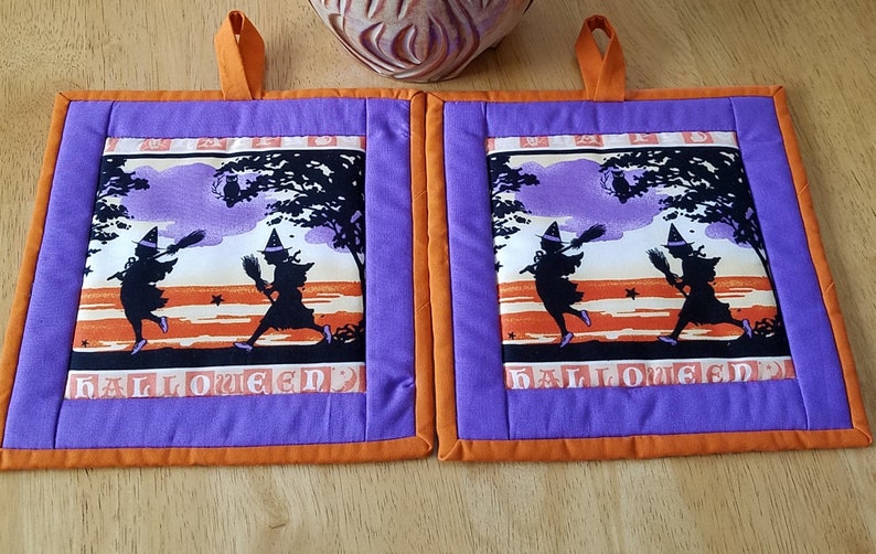 May include: Two purple and orange Halloween pot holders with a silhouette of two witches flying with broomsticks in front of a purple and orange sunset. The pot holders have the word "Halloween" printed on them.