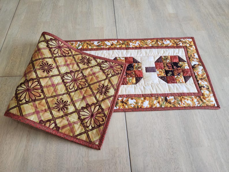 以下が含まれることがあります： キルト加工されたテーブルランナーと、お揃いのプレースマット。 ランナーは幾何学模様の中央のデザインと、秋の葉のボーダーが特徴です。 プレースマットは、黄色、赤、茶色の花柄とチェック柄です。