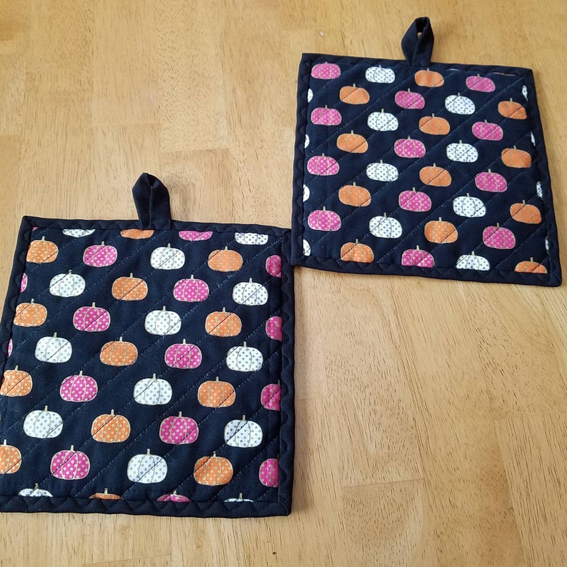May include: Two black quilted pot holders with a pumpkin pattern. The pumpkins are orange, white, and pink with white polka dots. The pot holders have a loop for hanging.
