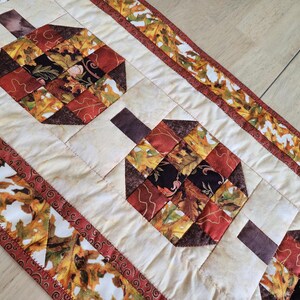 以下が含まれることがあります： 赤、オレンジ、茶色、黒の色合いの、カボチャの形をした秋をテーマにしたキルトテーブルランナー。 ランナーは、葉の模様のボーダーが付いたクリーム色の背景です。 秋の季節の装飾的なアクセント。