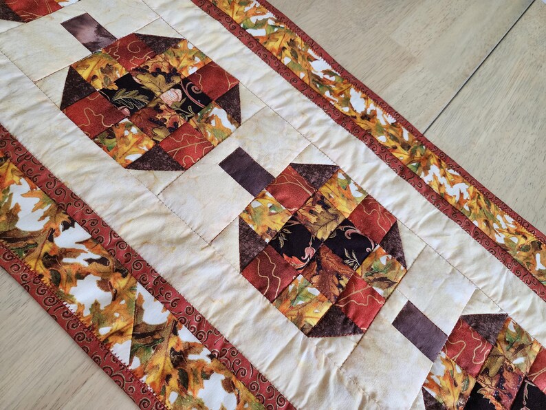 以下が含まれることがあります： 赤、オレンジ、茶色、黒の色合いの、カボチャの形をした秋をテーマにしたキルトテーブルランナー。 ランナーは、葉の模様のボーダーが付いたクリーム色の背景です。 秋の季節の装飾的なアクセント。