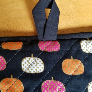 May include: Black quilted pot holder with a black fabric loop. The pot holder has a pattern of white, orange, and pink pumpkins with gold polka dots.