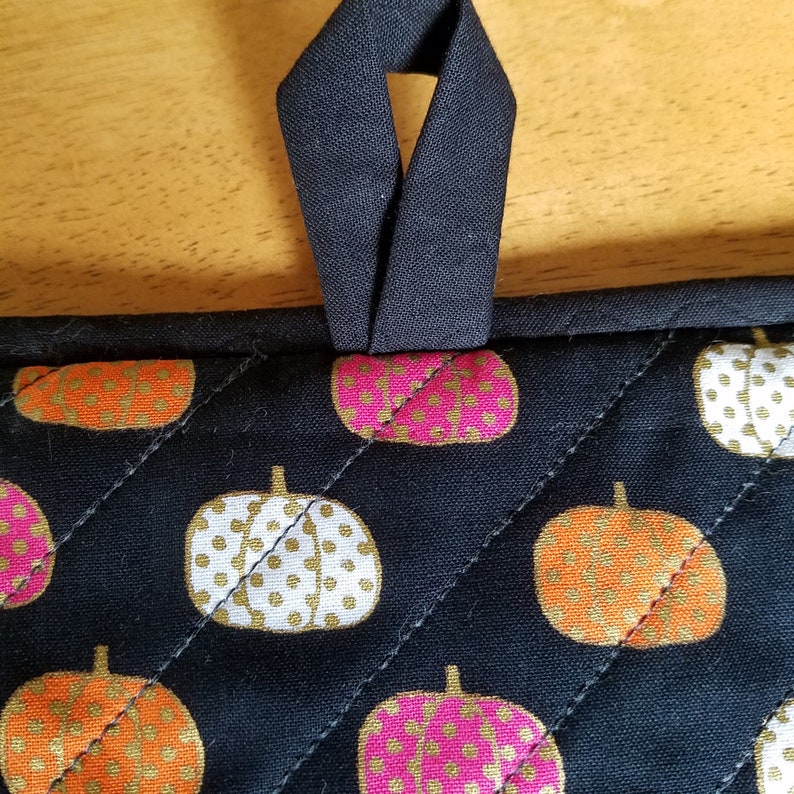 May include: Black quilted pot holder with a black fabric loop. The pot holder has a pattern of white, orange, and pink pumpkins with gold polka dots.