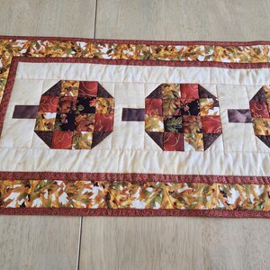 以下が含まれることがあります： 秋をテーマにした生地で作られた、クリーム色の中心部と3つの八角形のブロックが特徴のキルトテーブルランナー。茶色の長方形がブロックを接続しています。ボーダーには、秋の葉の繰り返し模様があります。