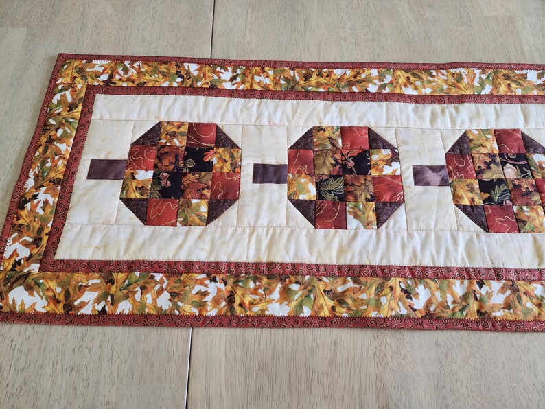 以下が含まれることがあります： 秋をテーマにした生地で作られた、クリーム色の中心部と3つの八角形のブロックが特徴のキルトテーブルランナー。茶色の長方形がブロックを接続しています。ボーダーには、秋の葉の繰り返し模様があります。