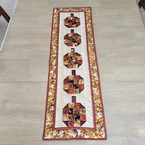 以下が含まれることがあります： 長方形のキルトテーブルランナー。カボチャのデザインが繰り返されています。カボチャは、オレンジ、茶色、赤の色合いのパッチワークの正方形で作られています。ランナーはクリーム色の背景と、秋の葉のボーダーが付いています。