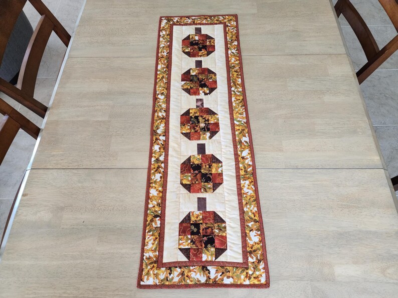 以下が含まれることがあります： 長方形のキルトテーブルランナー。カボチャのデザインが繰り返されています。カボチャは、オレンジ、茶色、赤の色合いのパッチワークの正方形で作られています。ランナーはクリーム色の背景と、秋の葉のボーダーが付いています。