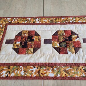 以下が含まれることがあります： 秋をテーマにしたキルトテーブルランナー。ランナーは、赤、オレンジ、茶色の秋の葉の幾何学的な形をしたクリーム色のセンターが特徴です。ボーダーは葉の模様と濃い赤色のトリムで飾られています。