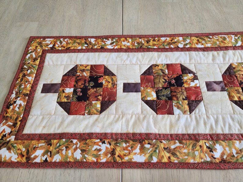 以下が含まれることがあります： 秋をテーマにしたキルトテーブルランナー。ランナーは、赤、オレンジ、茶色の秋の葉の幾何学的な形をしたクリーム色のセンターが特徴です。ボーダーは葉の模様と濃い赤色のトリムで飾られています。