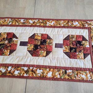 以下が含まれることがあります： 秋をテーマにしたキルトのテーブルランナー。ランナーは、赤、茶色、黒の色合いの3つの八角形の形をしたクリーム色のセンターを持っています。ボーダーは秋の葉で飾られています。