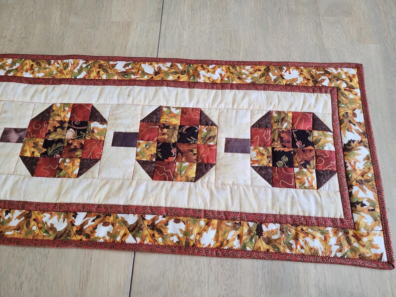 以下が含まれることがあります： 秋をテーマにしたキルトのテーブルランナー。ランナーは、赤、茶色、黒の色合いの3つの八角形の形をしたクリーム色のセンターを持っています。ボーダーは秋の葉で飾られています。