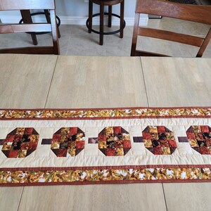 以下が含まれることがあります： クリーム色のセンターと秋の葉のボーダーが付いた、長方形のキルトテーブルランナー。中央には、赤、茶色、オレンジの色合いのパッチワークデザインの4つの八角形のブロックがあります。