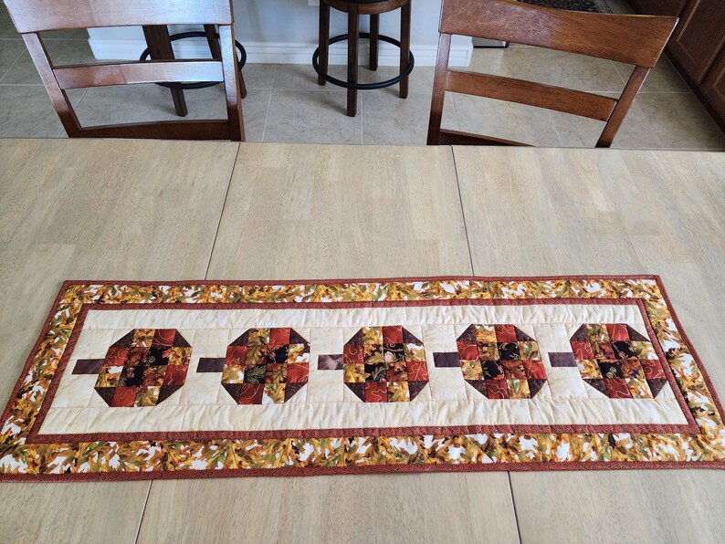 以下が含まれることがあります： クリーム色のセンターと秋の葉のボーダーが付いた、長方形のキルトテーブルランナー。中央には、赤、茶色、オレンジの色合いのパッチワークデザインの4つの八角形のブロックがあります。