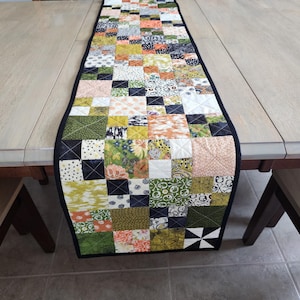 May include: A colorful patchwork table runner with a black border, displayed on a light wood table. The runner features a variety of square fabric patches in shades of green, orange, pink, and black. The table runner is approximately 60 inches long.