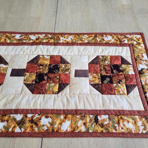 以下が含まれることがあります： 秋色の八角形の模様が繰り返されるキルトのテーブルランナー。 ランナーはクリーム色の中心に茶色のアクセントがあり、秋の葉のボーダーが付いています。 幅は約30cmです。