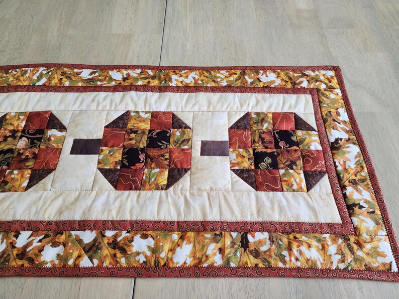 以下が含まれることがあります： 秋色の八角形の模様が繰り返されるキルトのテーブルランナー。 ランナーはクリーム色の中心に茶色のアクセントがあり、秋の葉のボーダーが付いています。 幅は約30cmです。