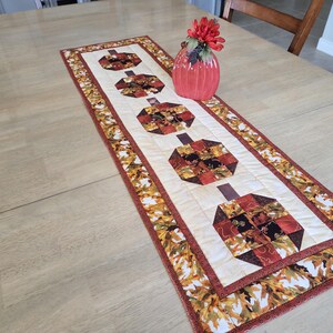 以下が含まれることがあります： クリーム色のセンターに4つのカボチャのデザインが施されたキルトテーブルランナー。カボチャはオレンジ、茶色、赤の色合いのパッチワーク生地で作られています。ボーダーには秋の葉の模様があります。赤い花瓶とフラワーアレンジメントがランナーの上にあります。