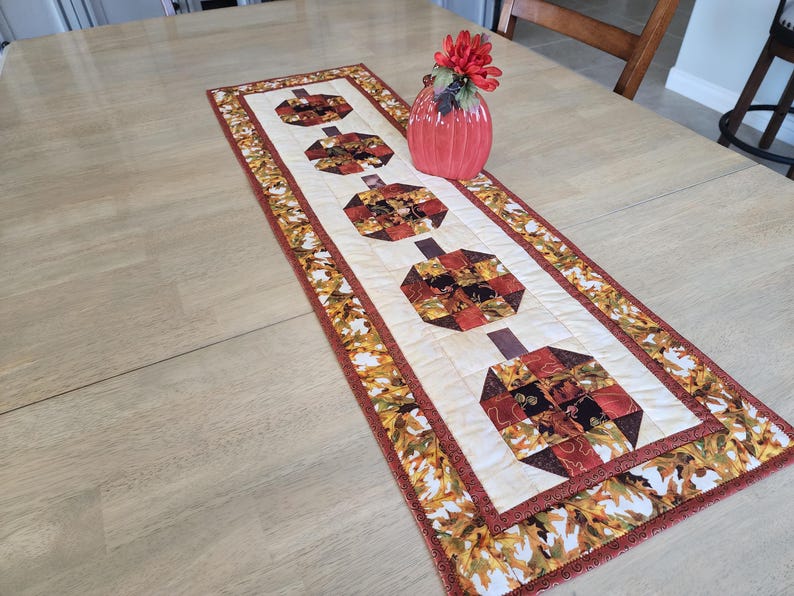 以下が含まれることがあります： クリーム色のセンターに4つのカボチャのデザインが施されたキルトテーブルランナー。カボチャはオレンジ、茶色、赤の色合いのパッチワーク生地で作られています。ボーダーには秋の葉の模様があります。赤い花瓶とフラワーアレンジメントがランナーの上にあります。