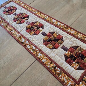 以下が含まれることがあります： 茶色、赤、オレンジの色合いで様式化されたカボチャの繰り返し模様が特徴のキルトテーブルランナー。カボチャはクリーム色の背景に配置され、秋の葉の模様で縁取られています。秋の装飾的なアクセント。