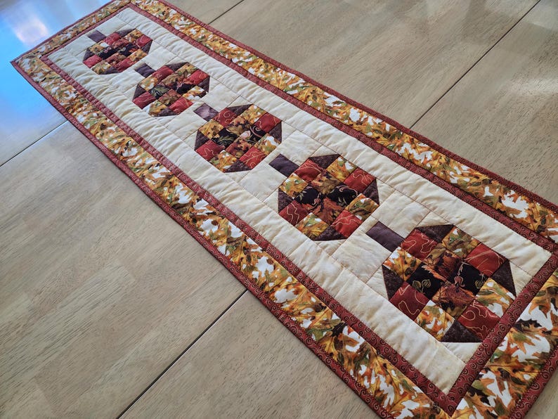 以下が含まれることがあります： 茶色、赤、オレンジの色合いで様式化されたカボチャの繰り返し模様が特徴のキルトテーブルランナー。カボチャはクリーム色の背景に配置され、秋の葉の模様で縁取られています。秋の装飾的なアクセント。