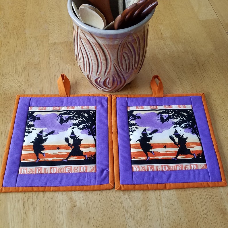 May include: Two purple and orange Halloween pot holders with a silhouette of two witches flying with broomsticks and the word "Halloween" printed on them.