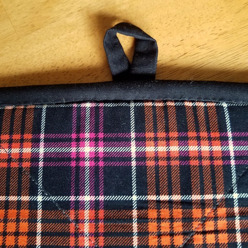 May include: Black pot holder with a plaid pattern in orange, pink, and white. The pot holder has a black loop handle.
