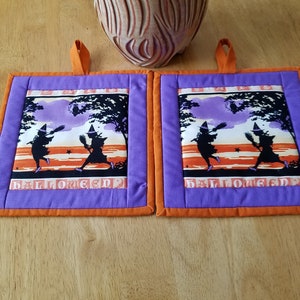 May include: Two purple and orange Halloween pot holders with a design of two witches flying on broomsticks with the text "HALLOWEEN" at the bottom.
