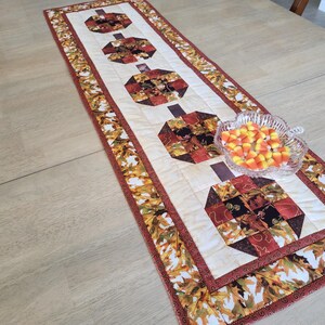 以下が含まれることがあります： クリーム色のセンターに4つのカボチャのデザインが施された、長方形の秋をテーマにしたテーブルランナー。カボチャは赤、茶色、黒の色合いのパッチワーク生地で作られています。ボーダーはオレンジ、黄色、茶色の葉の模様です。