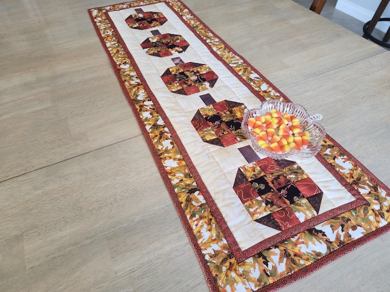 以下が含まれることがあります： クリーム色のセンターに4つのカボチャのデザインが施された、長方形の秋をテーマにしたテーブルランナー。カボチャは赤、茶色、黒の色合いのパッチワーク生地で作られています。ボーダーはオレンジ、黄色、茶色の葉の模様です。
