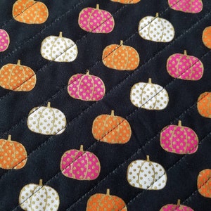May include: A black fabric with a repeating pattern of orange, white, and pink pumpkins with gold polka dots.