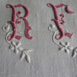 Peut inclure: Gros plan sur les initiales brodées 'R' et 'F' en fil rose sur un tissu blanc. Les lettres sont dans un style décoratif et cursif avec des accents floraux blancs. La broderie est détaillée et délicate.