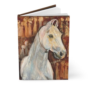 Pode incluir: Um diário de capa dura com um retrato de cavalo branco, lombada e páginas marrons. O cavalo tem um rosto detalhado com uma rédea escura e um fundo de figuras em tons de marrom e vermelho.