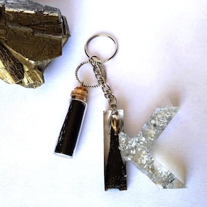 Peut inclure: Un porte-clés en argent avec un charme en forme de lettre K et un petit charme en verre en forme de fiole. Le charme en forme de lettre K est rempli de paillettes argentées et le charme en forme de fiole est rempli de paillettes noires.