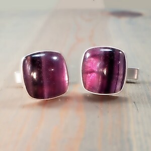 May include: A pair of cufflinks featuring square-shaped, deep purple, translucent stones with smooth, rounded edges. Each stone is set in a polished silver-toned metal frame, showcasing a rich, dark violet hue with subtle internal banding and light reflection.