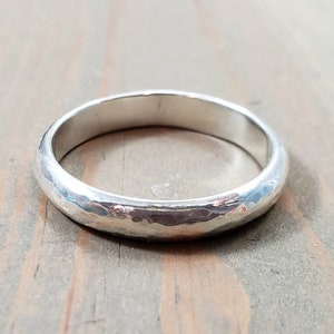 May include: A silver-coloured metal ring with a rounded profile and a textured, hammered surface finish. The band features a consistent width throughout its circular shape, reflecting light across its metallic exterior and polished inner edge.