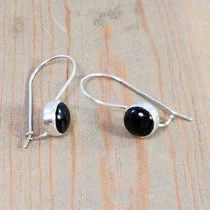 May include: A pair of silver-toned earrings with a black, oval-shaped stone set in a bezel. The earrings have a simple, hook-style design, and are displayed on a light-colored wooden surface. The earrings are a classic jewelry item.