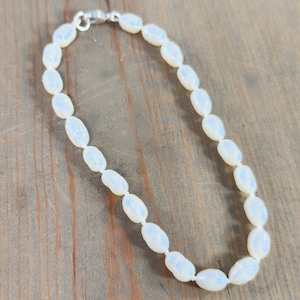 May include: An elegant bracelet composed of iridescent, oval beads. The beads display a pearlescent quality, with subtle blue and white reflections. The bracelet is fastened with a silver clasp. It is displayed on a wooden surface.