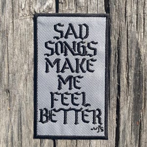May include: A gray embroidered patch with black text that reads "Sad Songs Make Me Feel Better".