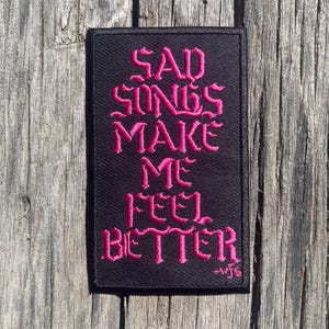 May include: Black embroidered patch with pink text that reads "SAD SONGS MAKE ME FEEL BETTER" with a small signature at the bottom.