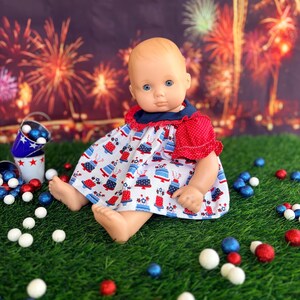 May include: A doll wearing a patriotic-themed dress with a red and white cake pattern, red polka dot sleeves, and a navy blue collar. The doll is surrounded by red, white, and blue decorative balls on a green surface, with a fireworks backdrop.