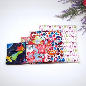 May include: Five reusable snack bags in different patterns. The bags are made of fabric and have a fold-over top with a snap closure. The patterns include a colorful floral design, a white background with pink giraffes, and a dark blue background with a colorful abstract design.