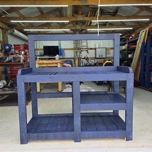 Puede incluir: Un gran banco de trabajo de madera azul con una superficie superior de listones, dos estantes inferiores y un tablero trasero. El banco está diseñado para tareas de jardinería al aire libre y tiene un aspecto robusto y rústico.