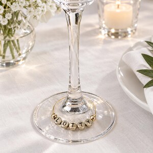 May include: A circular gold-toned wire wine glass charm featuring five small, round, metallic beads. Each bead is embossed with a black letter, collectively spelling the name LOTTY. The charm is positioned around the base of a clear glass stem.