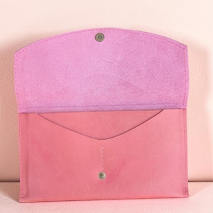 Könnte beinhalten: Eine rosa Leder-Umschlag-Clutch mit Klappenverschluss. Die Innenseite ist in einem helleren Rosaton gehalten. Die Clutch ist geöffnet und gibt den Blick ins Innere frei. Ein Metall-Druckknopf sichert die Klappe. Der Hintergrund ist einfarbig rosa.