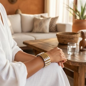Op de afbeelding: Een goud- en wit kralenarmband. De armband is een brede manchetstijl, met een herhalend geometrisch patroon. De kralen zijn goud en wit, wat een opvallend contrast creëert. De armband wordt om een pols gedragen.